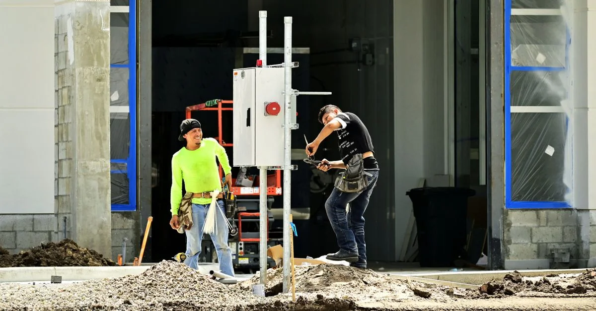Dos trabajadores de la construcción instalan un panel eléctrico fuera de un sitio de construcción, llevando equipo de seguridad.