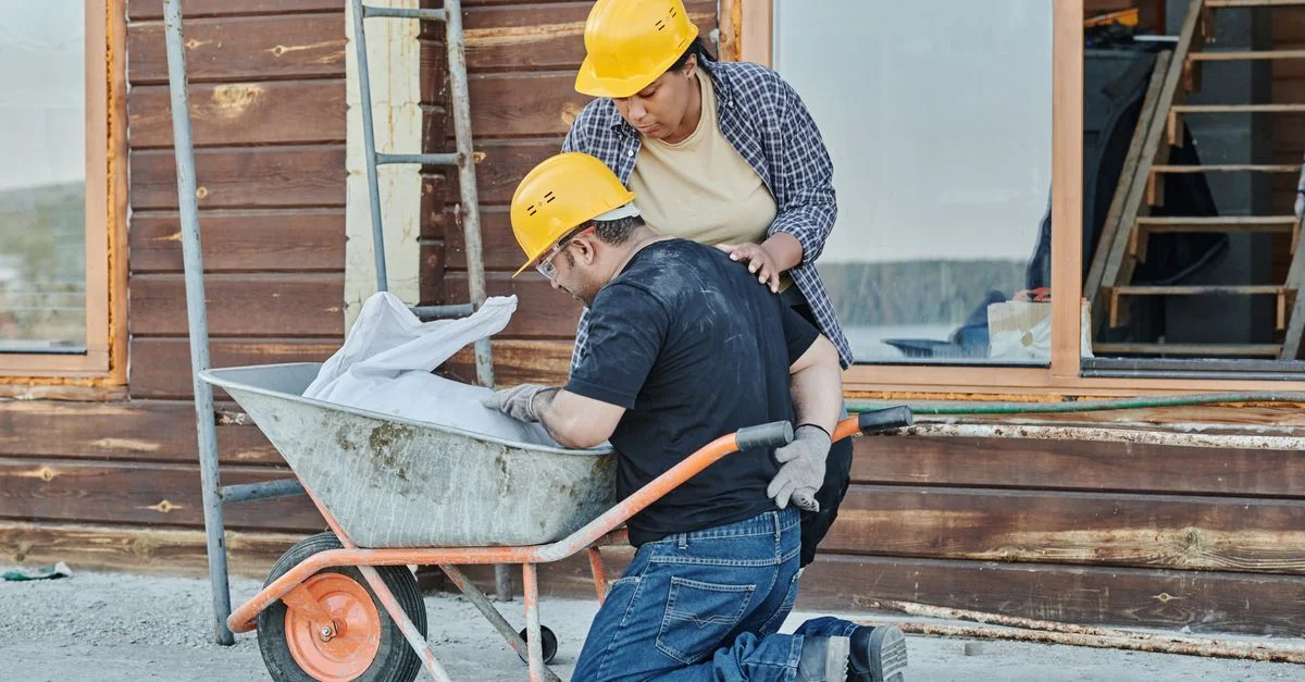 Un trabajador de la construcción recibiendo ayuda mientras está de rodillas con una carretilla en una obra.