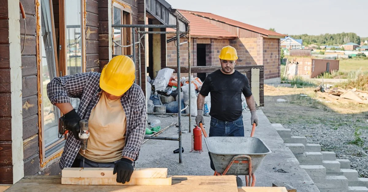 Trabajadores de la construcción construyendo una casa, usando una sierra y transportando materiales en una carretilla.
